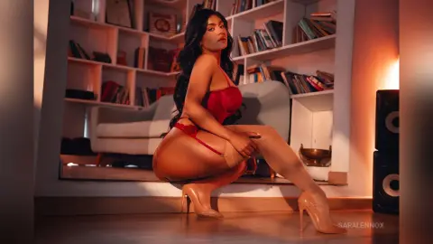 Glamorous woman in red lingerie and nude heels posing in a cozy, sunlit living room with bookshelves.