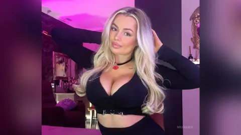 Blonde woman in a black crop top posing in a room with moody pink lighting.
