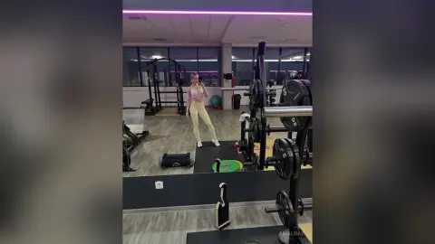 Fit woman in a pink sports bra and cream leggings taking a mirror selfie in a modern gym setting.