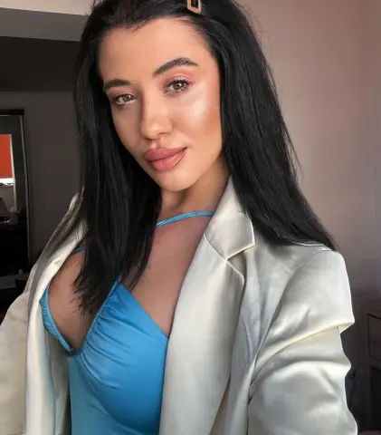 Young woman with long dark hair wearing a blue satin top and white blazer in a bright indoor setting.