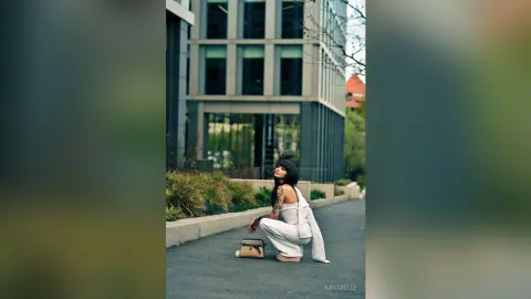 Stylish woman in white outfit squatting on a city sidewalk near a modern building with a chic, urban mood.