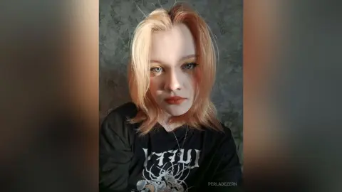 Blonde woman in a black graphic t-shirt posing with a moody expression against a floral wallpaper background.