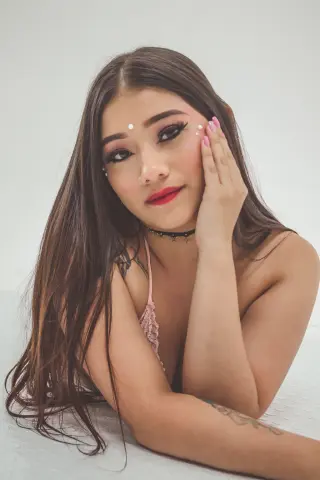 Young woman with long brown hair and pink makeup wearing a lace bikini in a bright, minimalist setting.