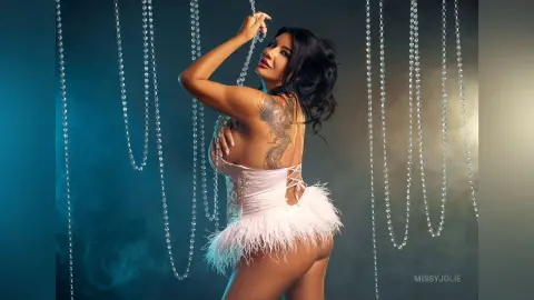 MissyJolie poses sideways in a white lace corset and feathered skirt, showing her bare back and side-boob against a dark, smoky background.