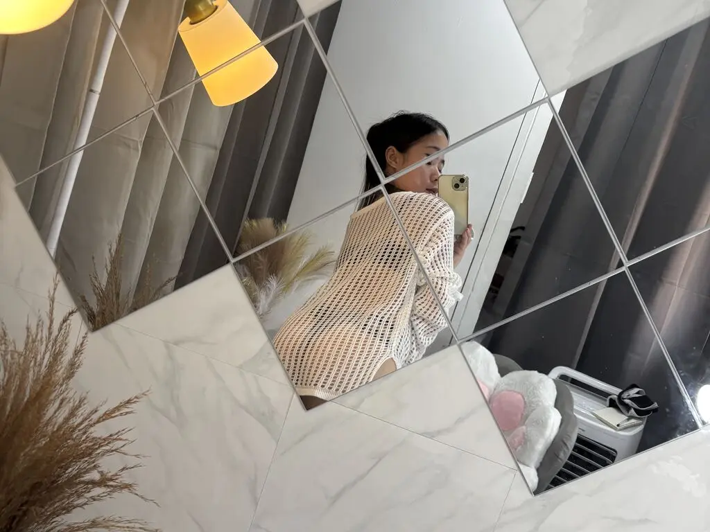 Young woman in a white knit sweater taking a mirror selfie in a modern, bright room.