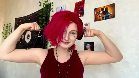 Smiling woman with bright pink hair in a maroon tank top flexing her muscles in a cozy bedroom setting.