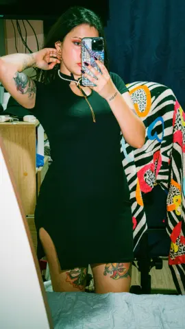 Young woman with tattoos wearing a black mini dress taking a mirror selfie in a cozy bedroom setting.