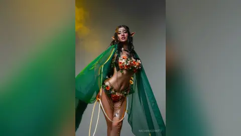 Ethereal forest elf cosplayer in a floral green outfit and flowing cape against a misty, magical background.