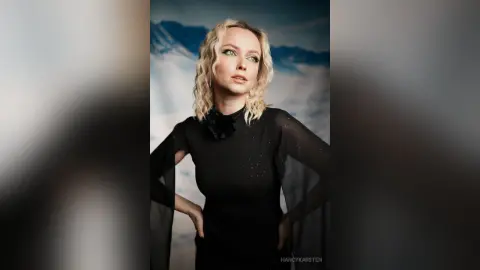 Blonde woman in a black sheer long-sleeve top posing against a dreamy mountain landscape background.