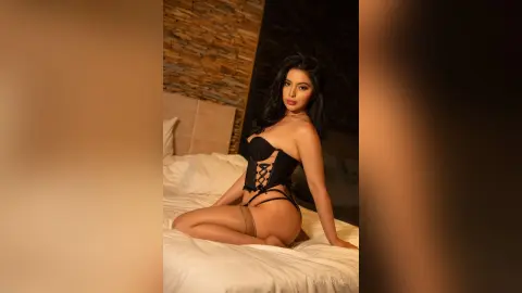 VickyPortman poses on a white bed wearing a black lace corset and black thong, showing her bare back and legs.