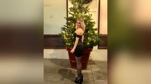 Elegant woman in a black cocktail dress posing by a glowing Christmas tree in a festive indoor setting.