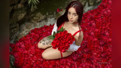Young woman in red lingerie holding a rose bouquet while sitting on red rose petals in a romantic garden setting.