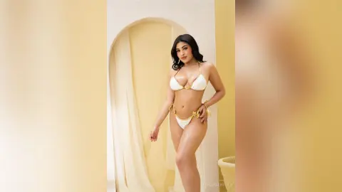 Glamorous woman in a white bikini posing in a sunlit, minimalist Mediterranean-style room.