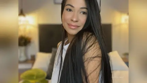 Smiling woman with long dark hair wearing a white tank top in a cozy, brightly lit bedroom setting.