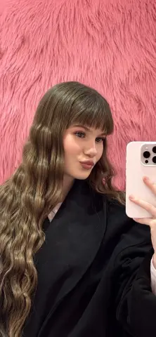 AshleyFierce poses for a mirror selfie wearing a black coat, showing off her long wavy brown hair and bangs against a pink furry background.