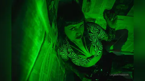 Moody portrait of a woman in a zebra print top posing in a green lit room with newspaper decor.