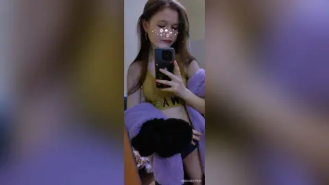 Young woman in a yellow crop top and purple cardigan taking a mirror selfie in a cozy indoor setting.