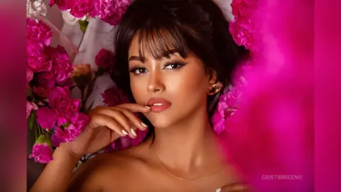 CristiBriceno poses with her hand near her lips, wearing a gold necklace and earrings, surrounded by bright pink flowers.