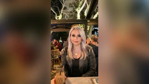 Smiling blonde woman in a black leather jacket enjoying a drink at an outdoor evening restaurant terrace.