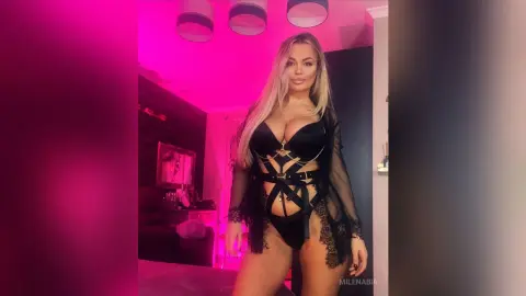 Blonde woman in black lace lingerie and sheer robe posing in a pink neon lit bedroom.