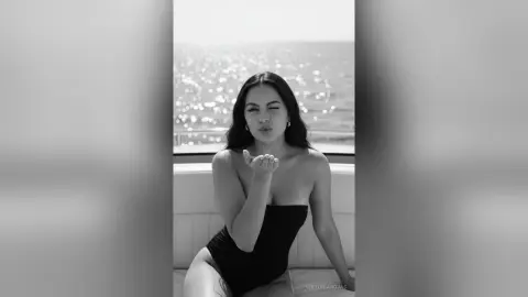 Winking woman in a black bodysuit sitting on a boat against a sparkling ocean background.