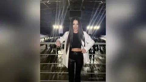 LeonidaAbdon poses in a white blazer and black leather pants, standing against a dark, lit stage background with long dark hair.
