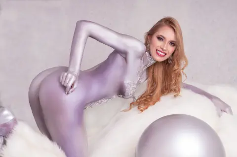 Smiling blonde woman in a shimmering silver bodysuit posing on white faux fur in a bright studio setting.