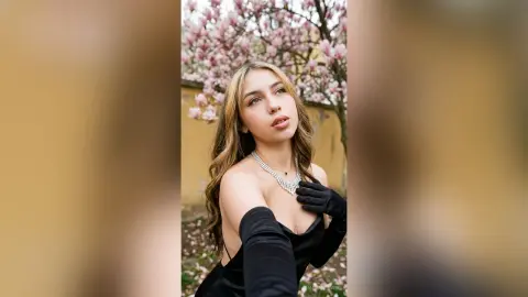 Elegant woman in a black dress and gloves posing under blooming pink flowers in a dreamy garden setting.