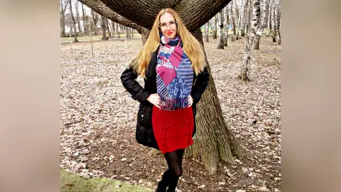 Blonde woman in a black coat, red skirt, and patterned scarf standing by a large tree in a fall forest.