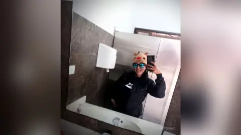 Smiling woman in a black jacket and funny animal hat taking a mirror selfie in a modern bathroom.