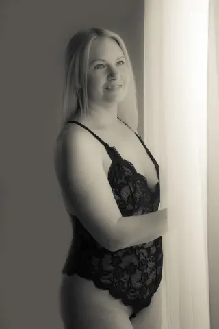 Ember_Sinn poses in a black lace bodysuit, looking over her shoulder while standing near a bright window.