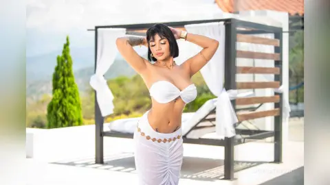 Stylish woman in a white bikini top and skirt posing under a sunny outdoor cabana with a tropical view.