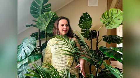 DariaCalm poses behind large green monstera leaves, wearing a light yellow knit tank top with a floral detail.