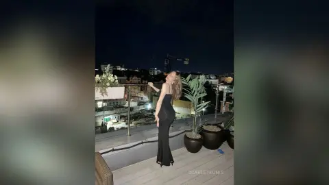 Elegant woman in a black evening gown posing on a rooftop balcony at night with city lights in the background.