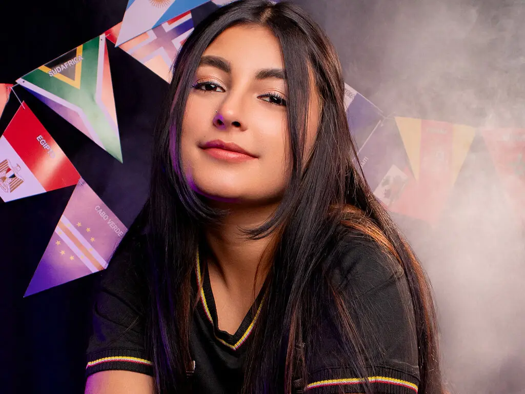 KylieThomps poses with long dark hair wearing a black v-neck shirt against a background of colorful flags.