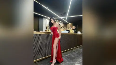 CristalMatthews stands in a red strapless slit dress, showing off her long bare leg in a dimly lit room.