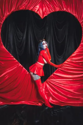 KimLennoxc poses in a red outfit and tall boots, framed by a large red heart-shaped curtain against a black background.