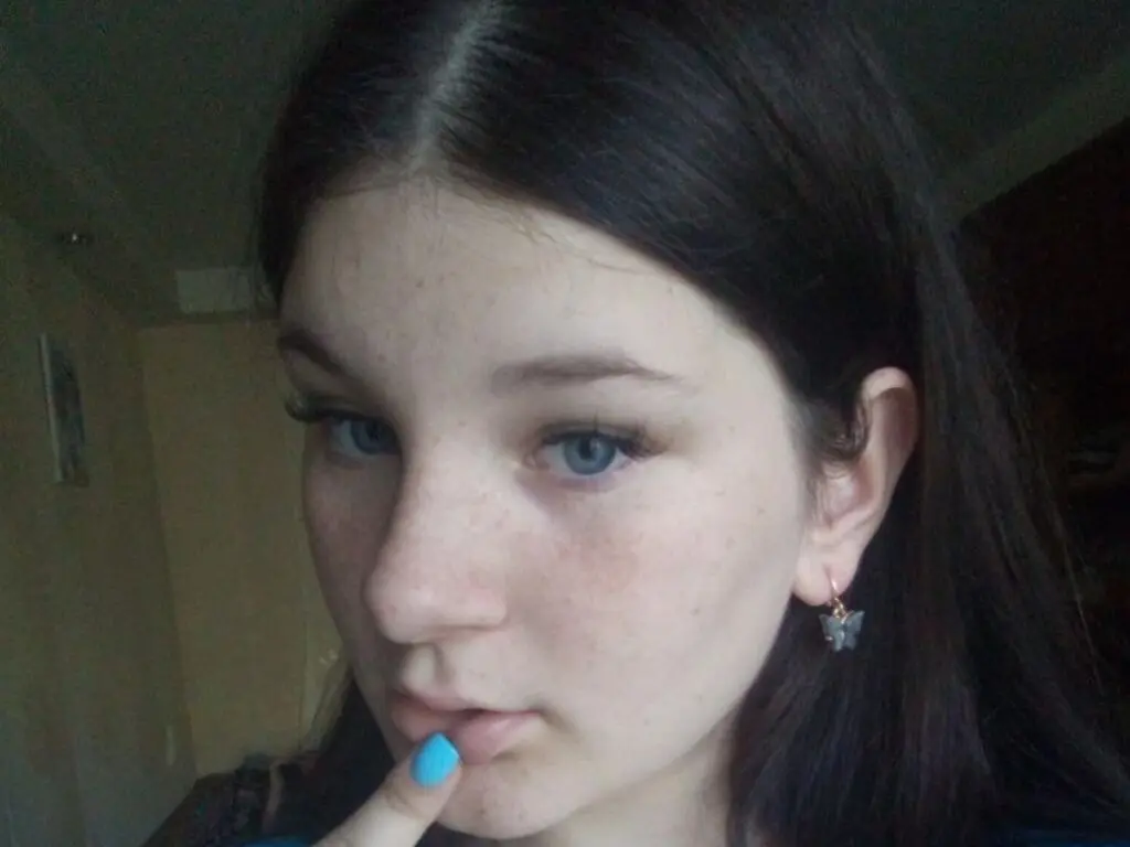 Close-up portrait of a young woman with freckles and long dark hair looking into the camera indoors.