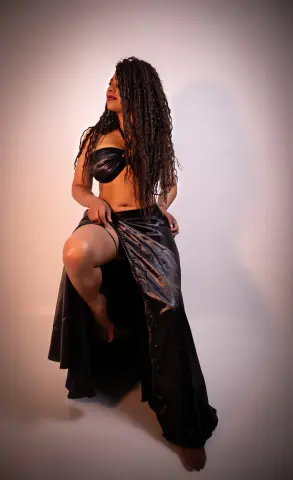 Woman with long curly hair in a black satin outfit posing against a neutral studio background.