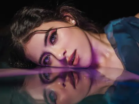 Close-up of a woman with glamorous eye makeup lying on a reflective surface in moody, purple lighting.