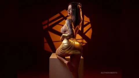 Glamorous woman in gold metallic outfit and jewelry posing against warm dramatic lighting in a studio setting.