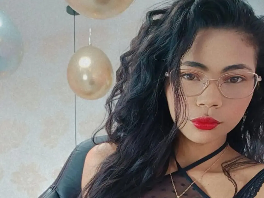 Young woman with curly dark hair and glasses wearing a black halter top in a festive room with gold balloons.