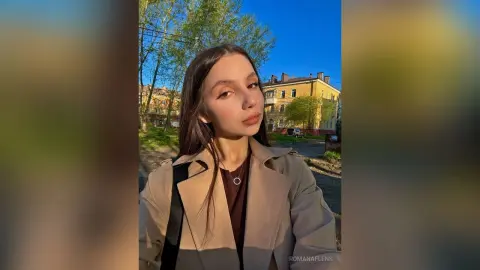 Young woman in a beige trench coat smiling outdoors in a sunny urban park setting.