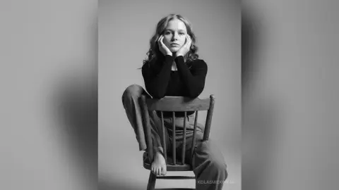 Melancholic young woman in a black turtleneck sitting on a wooden chair against a gray studio background.
