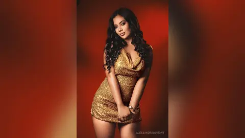 AlessandraDumont poses in a tight, gold sequin mini dress against a red background, showing off her long, wavy dark hair.