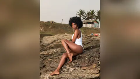 Beautiful Black woman in a white bodysuit relaxing on coastal rocks under a soft sunset sky.