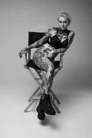 Tattooed woman in black lingerie and combat boots sitting on a director's chair in a moody black and white portrait.