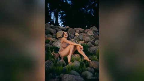 Elegant woman in a tan dress sitting on a rocky landscape under a moody forest sky.