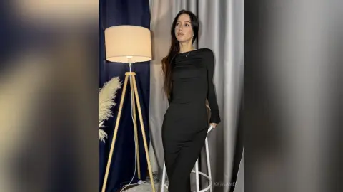 Elegant woman in a sleek black long-sleeve dress standing by a tripod floor lamp in a modern room.