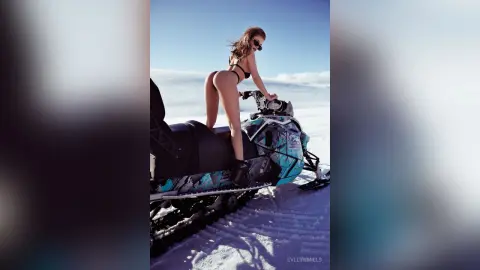 EvelynMills poses naked on a blue snowmobile in a bright snowy landscape, wearing sunglasses and looking over her shoulder.